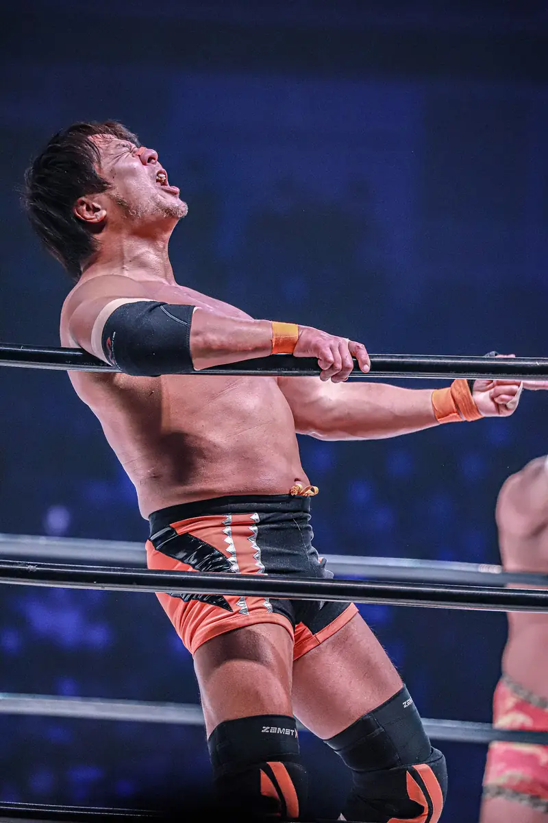Satoshi Kojima in his orange gear, arm stretched out along the ropes