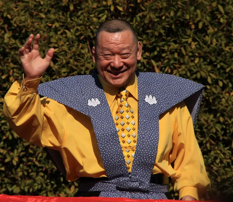 Animal Hamaguchi smiling and waving, in a yellow and blue outfit