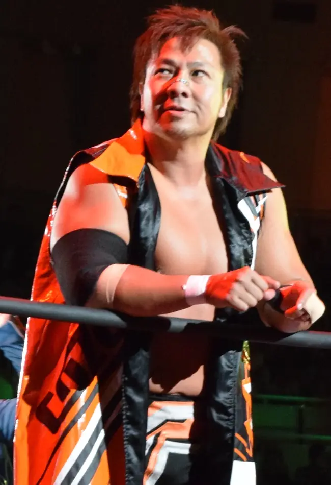 Kojima in an orange and black robe, leaning on the ropes